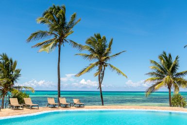 Ein strahlend blauer Himmel über einem Pool, umgeben von Palmen und dem glitzernden Wasser des Ozeans im Hintergrund.