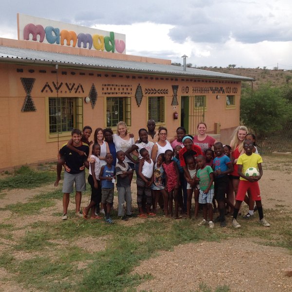Eine Gruppe von Kindern und Erwachsenen posiert fröhlich vor einem bunten Gebäude mit dem Schriftzug 'mammadu'.