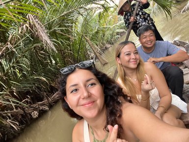 Drei Personen lächeln fröhlich auf einem Boot im Mekong-Delta, umgeben von üppigem, grünem Pflanzenbewuchs.