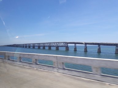 Eine lange Brücke erstreckt sich über das glitzernde Wasser, unter einem strahlend blauen Himmel mit wenigen Wolken.