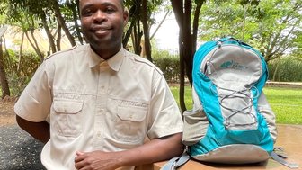 Ein lächelnder Mann in khakifarbener Kleidung steht neben einem Rucksack auf einem Tisch aus Stein in einer grünen Umgebung.