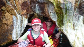 Ein Junge und eine Frau paddeln in einem Kajak durch eine enge Höhle, umgeben von bunten Felsen und Wasser.