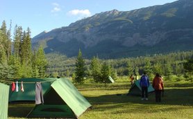 Zwei Personen stehen auf einem Campingplatz mit grünen Zelten, umgeben von hohen Bäumen und majestätischen Bergen im Hintergrund.