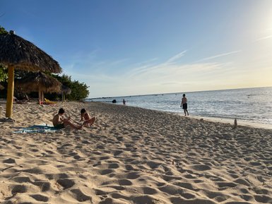 Zwei Kinder sitzen im Sand am Strand, während ein Junge im Hintergrund am Wasser steht und die Wellen beobachtet.