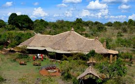 Eine luftige Ansicht eines traditionellen, strohgedeckten Camps inmitten einer grünen, bewaldeten Landschaft unter blauem Himmel.