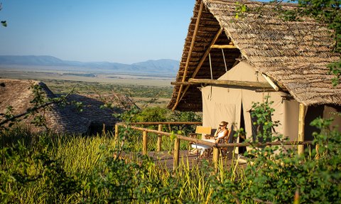 Eine Frau sitzt entspannt auf der Veranda eines Zeltes und genießt die Aussicht auf die weite Landschaft des Ngorongoro-Kraters.