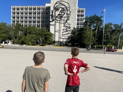 Zwei Jungen stehen auf einem Platz und betrachten ein großes Wandbild von Che Guevara auf einem Gebäude im Hintergrund.