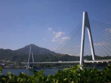 Eine moderne Hängebrücke erstreckt sich über einen ruhigen Fluss, umgeben von sanften Hügeln und klarem Himmel.