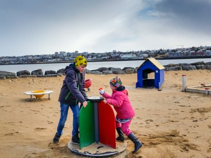 Zwei Kinder spielen auf einem Sandspielplatz, während im Hintergrund eine Stadtlandschaft und ein ruhiger Wasserbereich sichtbar sind.
