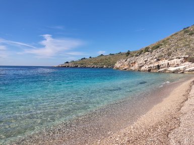 Ein ruhiger Strand mit klarem, türkisfarbenem Wasser und sanften Wellen, umgeben von grünen Hügeln und blauen Himmel.