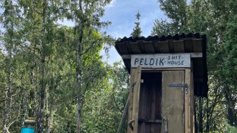 Eine rustikale Holzhütte mit der Aufschrift 'PELDIK SHIT HOUSE' steht umgeben von hohen Bäumen und dichtem Grün.