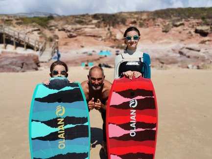 Drei Personen stehen am Strand, halten bunte Bodyboards und lächeln fröhlich in die Kamera, umgeben von einer schönen Landschaft.
