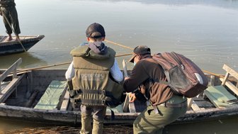 Ein Junge in einer Schwimmweste beobachtet einen Mann, der ein Boot am Ufer eines ruhigen Flusses vorbereitet.