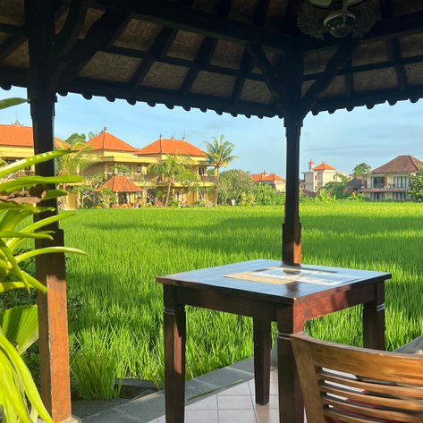 Ein gemütlicher Pavillon mit einem Holztisch und einem Stuhl, umgeben von üppigen Reisfeldern und balinesischer Architektur.