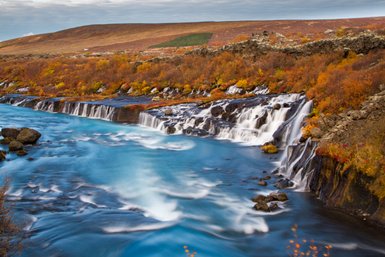 Ein malerischer Wasserfall fließt über schwarze Felsen, umgeben von buntem Herbstlaub und klarem, blauem Wasser.