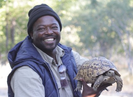 Ein lächelnder Mann hält eine Schildkröte in der Natur, umgeben von Bäumen und sanftem Licht.
