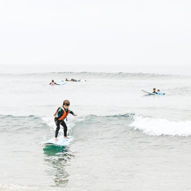 Ein Junge surft auf einer Welle, während andere Surfer im Hintergrund auf ihren Brettern paddeln.