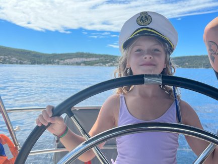 Ein fröhliches Kind mit einem Kapitänshut steuert ein Segelboot unter einem strahlend blauen Himmel.