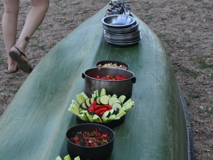 Ein Kanu liegt am Ufer, während frische Salate und Snacks in Schalen auf dem Boot angerichtet sind.
