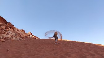 Eine Person steht auf einer sandigen Düne und wirbelt Sand in die Luft, während die Sonne am Horizont scheint.