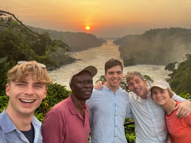 Fünf Personen posieren fröhlich vor einer atemberaubenden Landschaft mit einem Sonnenuntergang über einem Fluss.