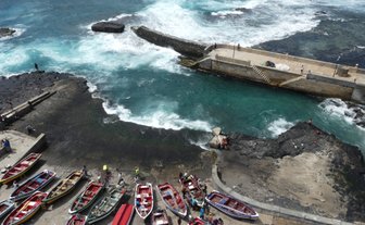 Bunte Fischerboote sind am Ufer eines lebhaften Wassers mit Wellen angedockt, während Menschen Aktivitäten im Hintergrund unternehmen.