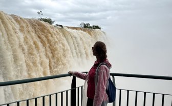 Eine Person in rot-weißer Jacke steht an einem Geländer und schaut fasziniert auf die mächtigen Wasserfälle.