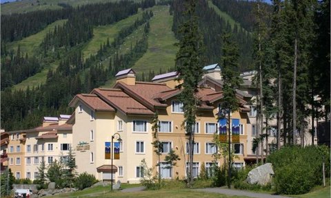 Die Außenansicht des Cahilty Lodge zeigt ein helles Gebäude, umgeben von hohen Bäumen und einer malerischen Berglandschaft.