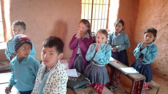 Sechs Kinder in blauen Hemden stehen in einem Klassenzimmer mit braunen Wänden und Holzbänken, einige beten.