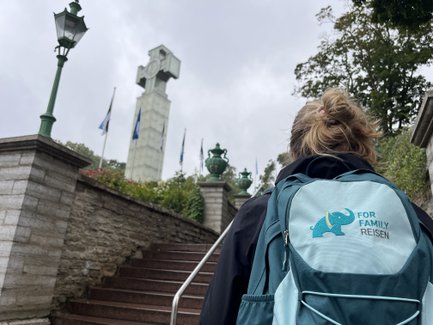 Eine Person mit einem Rucksack geht eine Treppe hinauf, während im Hintergrund ein hoher, markanter Turm sichtbar ist.