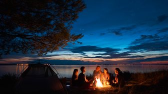 Eine Gruppe von fünf Freunden sitzt um ein Lagerfeuer am Ufer, während der Sonnenuntergang den Himmel in warmen Farben erleuchtet.