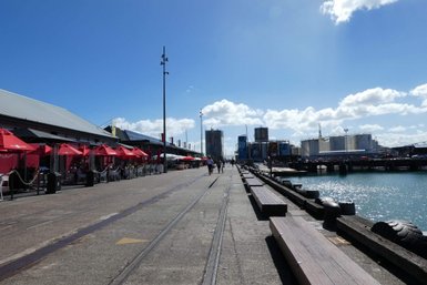 Ein belebter Hafen mit roten Sonnenschirmen, wo Menschen entlang der Promenade spazieren und die Sonne genießen.