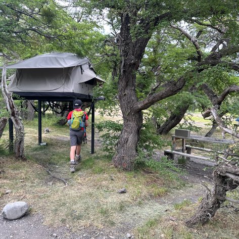 Ein Wanderer nähert sich einem erhöhten Zelt, umgeben von üppigem Grün und Bäumen in einer ruhigen Campingumgebung.