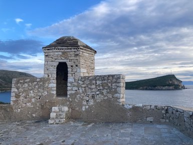 Eine alte Steinhütte mit einem gewölbten Dach steht auf einer Mauer, die über das Wasser und die umliegenden Hügel blickt.
