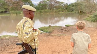 Ein Sicherheitsbeamter in Uniform beobachtet einen Jungen, der fasziniert auf einen Krokodil am Ufer schaut.