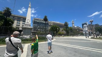 Ein Mann und ein Junge zeigen auf ein Denkmal in Buenos Aires, umgeben von modernen Gebäuden und Palmen unter blauem Himmel.