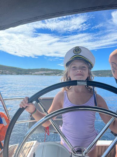 Ein fröhliches Kind mit einem Kapitänshut steuert ein Segelboot unter einem strahlend blauen Himmel.