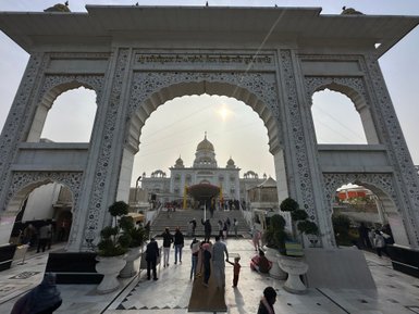 Ein majestätisches Tor mit kunstvollen Verzierungen führt zu einem beeindruckenden Gurudwara, umgeben von Besuchern.