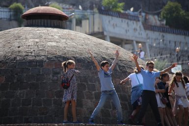 Eine Gruppe von Kindern spielt fröhlich vor einem großen, runden Gebäude aus Stein, das im Hintergrund sichtbar ist.