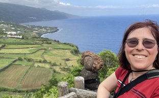 Eine Person mit Sonnenbrille lächelt vor einer atemberaubenden Küstenlandschaft mit grünem Terrain und blauem Meer.