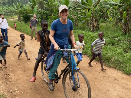 Eine Frau fährt auf einem Fahrrad, während Kinder fröhlich auf einem staubigen Weg hinterherlaufen und lachen.