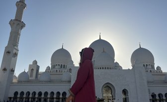 Eine Person in einem roten Gewand steht vor der beeindruckenden Sheikh Zayed Moschee, umgeben von majestätischen Kuppeln.