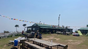 Eine Gruppe von Menschen sitzt an einem Tisch im Freien, umgeben von bunten Gebetsfahnen und einem klaren blauen Himmel.