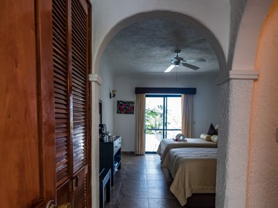 Ein einladender Blick in ein helles Zimmer mit zwei Betten, einem Ventilator und einem großen Fenster, das auf die Terrasse führt.