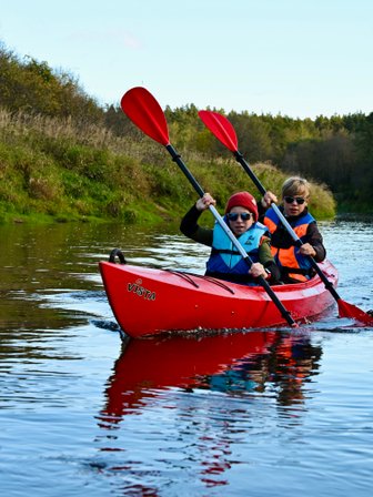 Zwei Kinder paddeln in einem roten Kajak auf einem ruhigen Fluss, umgeben von grünen Ufern und Bäumen im Hintergrund.