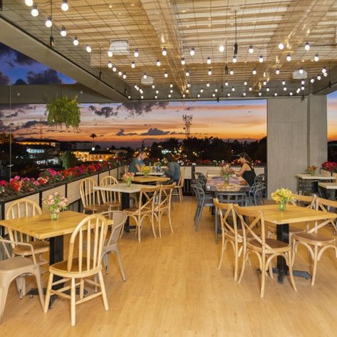 Eine stilvolle Terrasse mit Holztischen und Stühlen, geschmückt mit bunten Blumen und Lichtern, bietet einen atemberaubenden Sonnenuntergang.