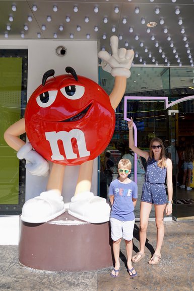 Ein großer, roter M&M's Charakter steht vor einem Geschäft, während ein Junge und ein Mädchen lächeln und posieren.