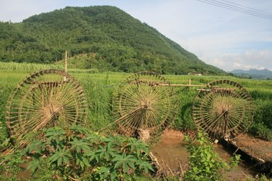 Drei große, hölzerne Wasserräder stehen in einer grünen Landschaft, umgeben von üppigem Gras und Bergen im Hintergrund.