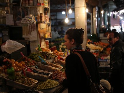Eine Frau betrachtet sorgfältig die bunten Obststände auf einem Markt, während ein Mann im Hintergrund zuschaut.