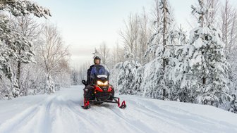 Ein Fahrer auf einem Schneemobil fährt durch eine verschneite Winterlandschaft, umgeben von schneebedeckten Bäumen.